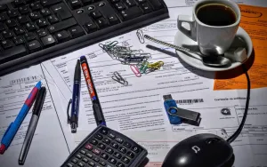 Cup of tea, keyboard, mouse, biros, calculator on top of papers.