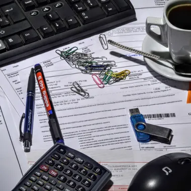 Cup of tea, keyboard, mouse, biros, calculator on top of papers.